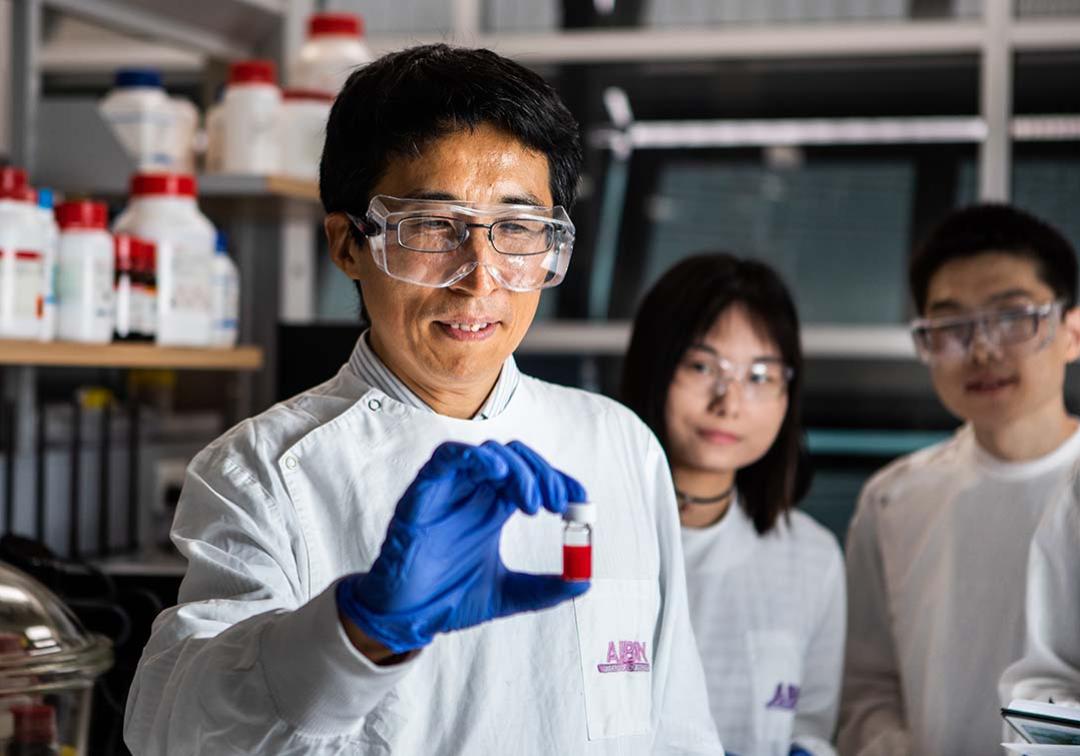 Man in lab coat and goggles holding vial. Two people wearing goggles in background looking at man. 