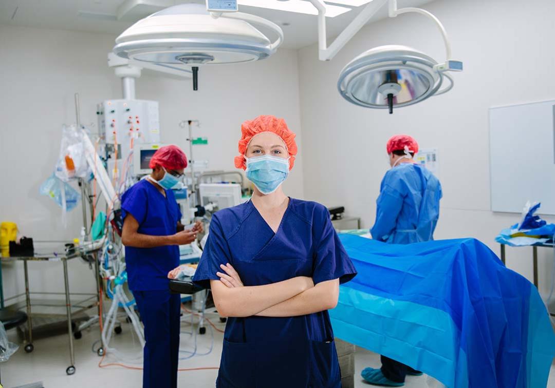 Woman in scrubs with crossed arms, smiling at camera. Medical professionals in scrubs and patient in the background.