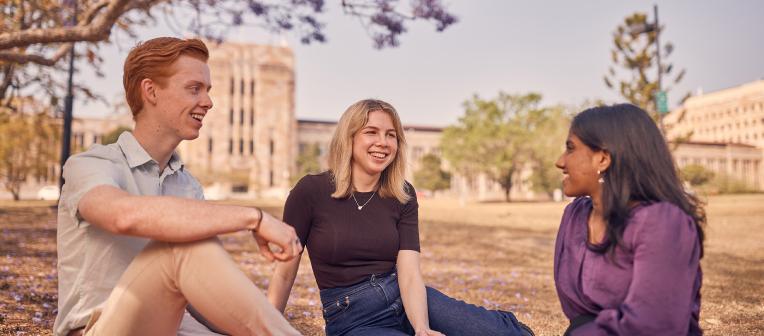 Three students sitting on the lawn at UQ with the Forgan Smith Building behind them.