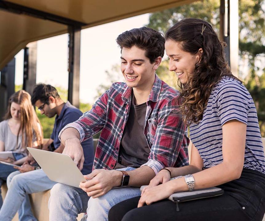 Two students looking at a computer, sitting in the UQ walkway area. Other students in background. 