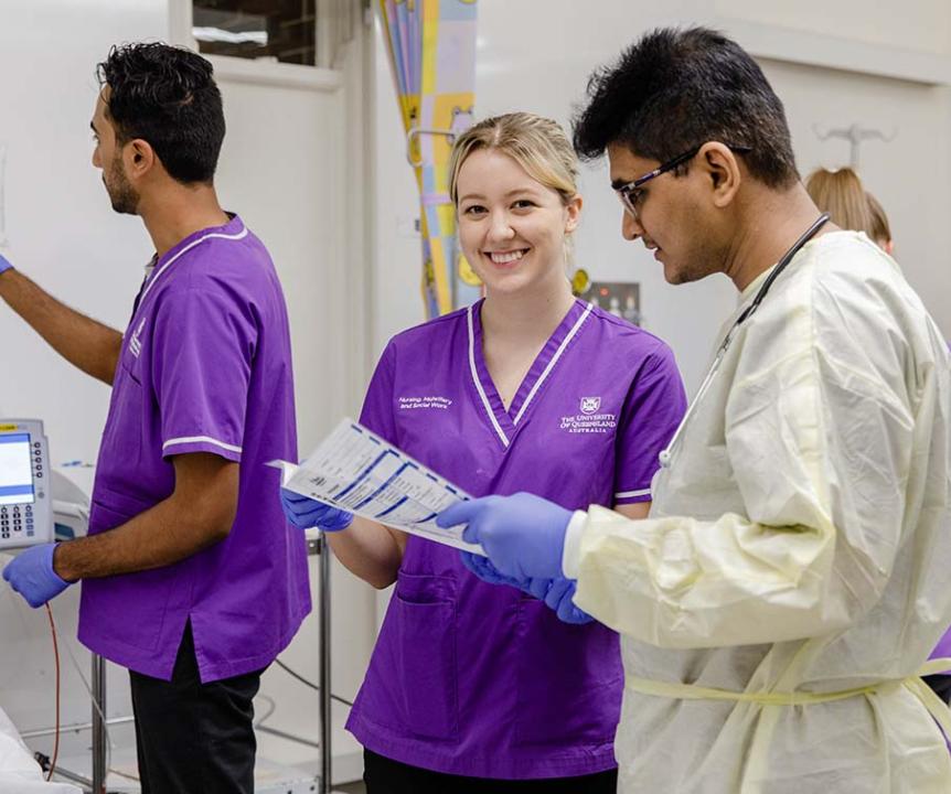 Woman in purple scrubs smiling at camera while being shown a medical document. Man in purple scrubs attending to IV in the background. 