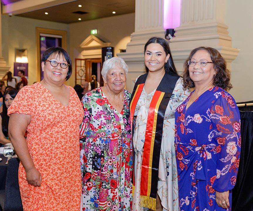 Four women stand smiling for the camera. One of the women is wearing a black, red and yellow sash.