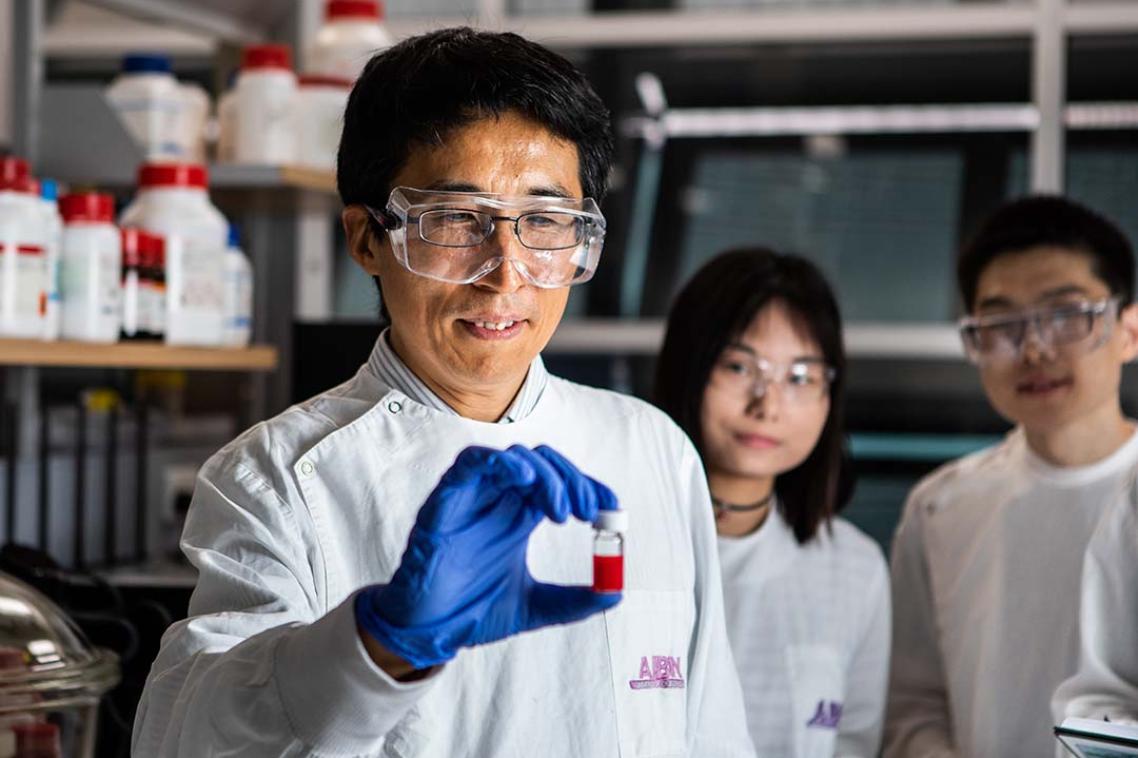 Man in lab coat and goggles holding vial. Two people wearing goggles in background looking at man. 