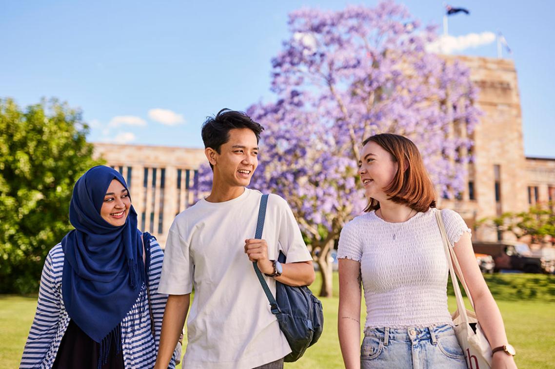 Three people walking together. A jacaranda tree and the sandstone Forgan Smith building appear in the background.